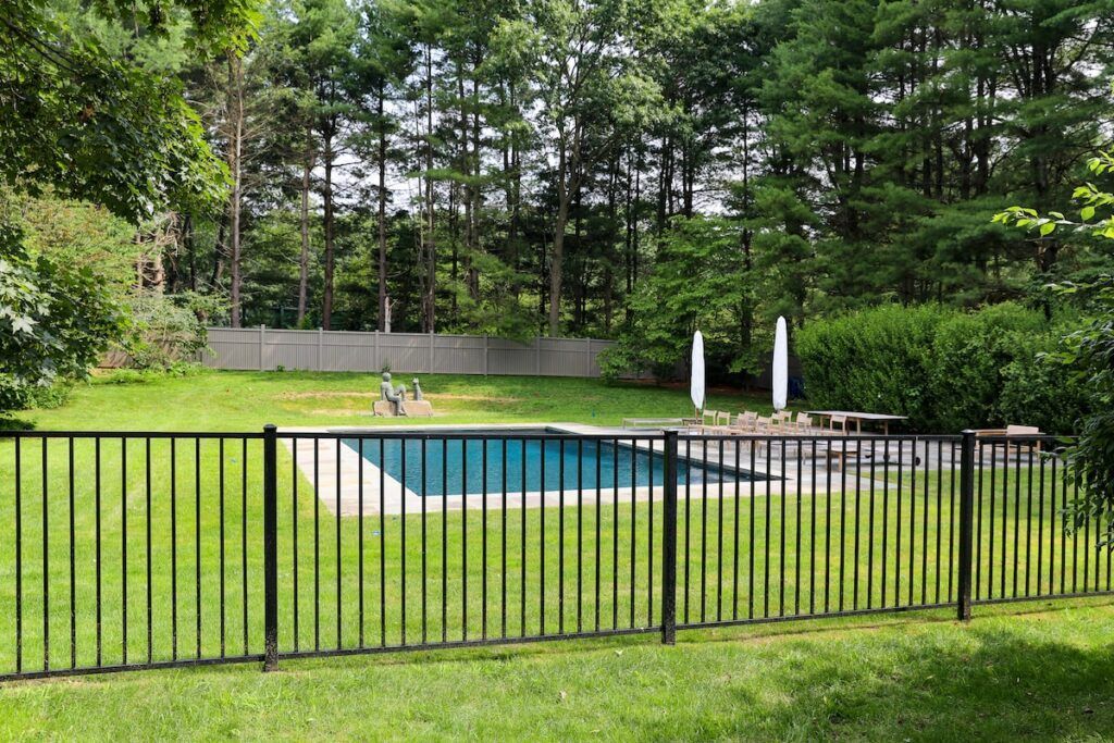 Black fence surrounds a backyard with a pool, grassy lawn, and tall trees.