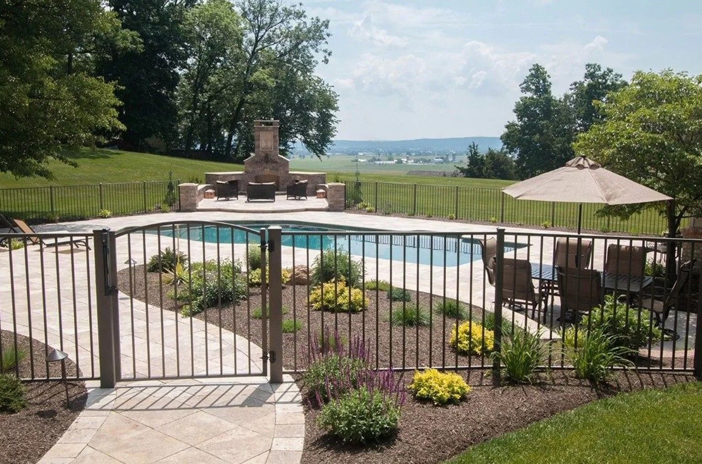 Outdoor pool with a wrought-iron fence, landscaping, and a stone fireplace with a distant view.