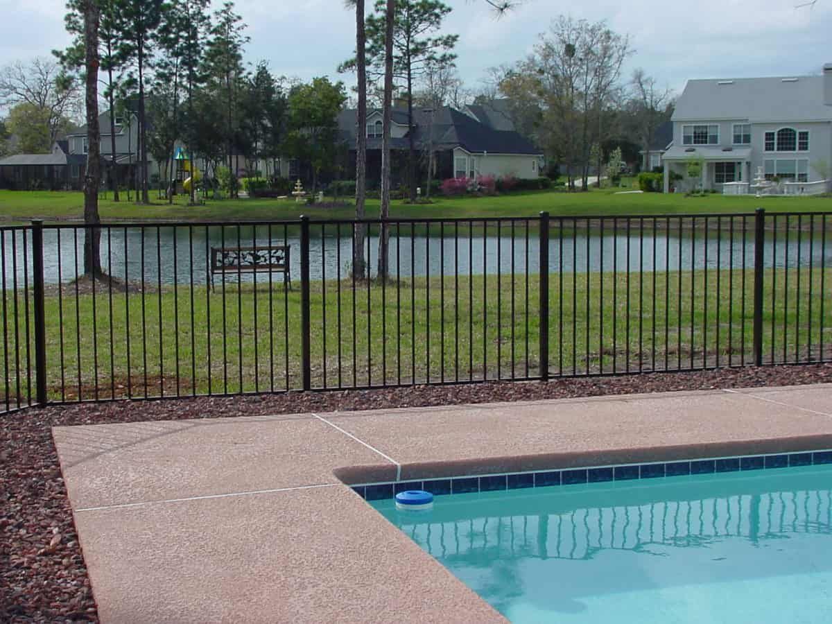 Black fence bordering a pool, overlooking a lake with houses in the distance.