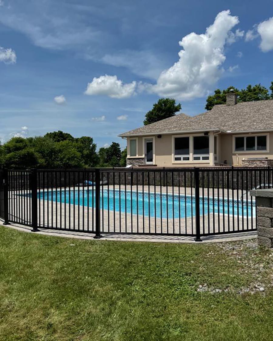 Black fence surrounds a swimming pool next to a beige house, under a cloudy blue sky.