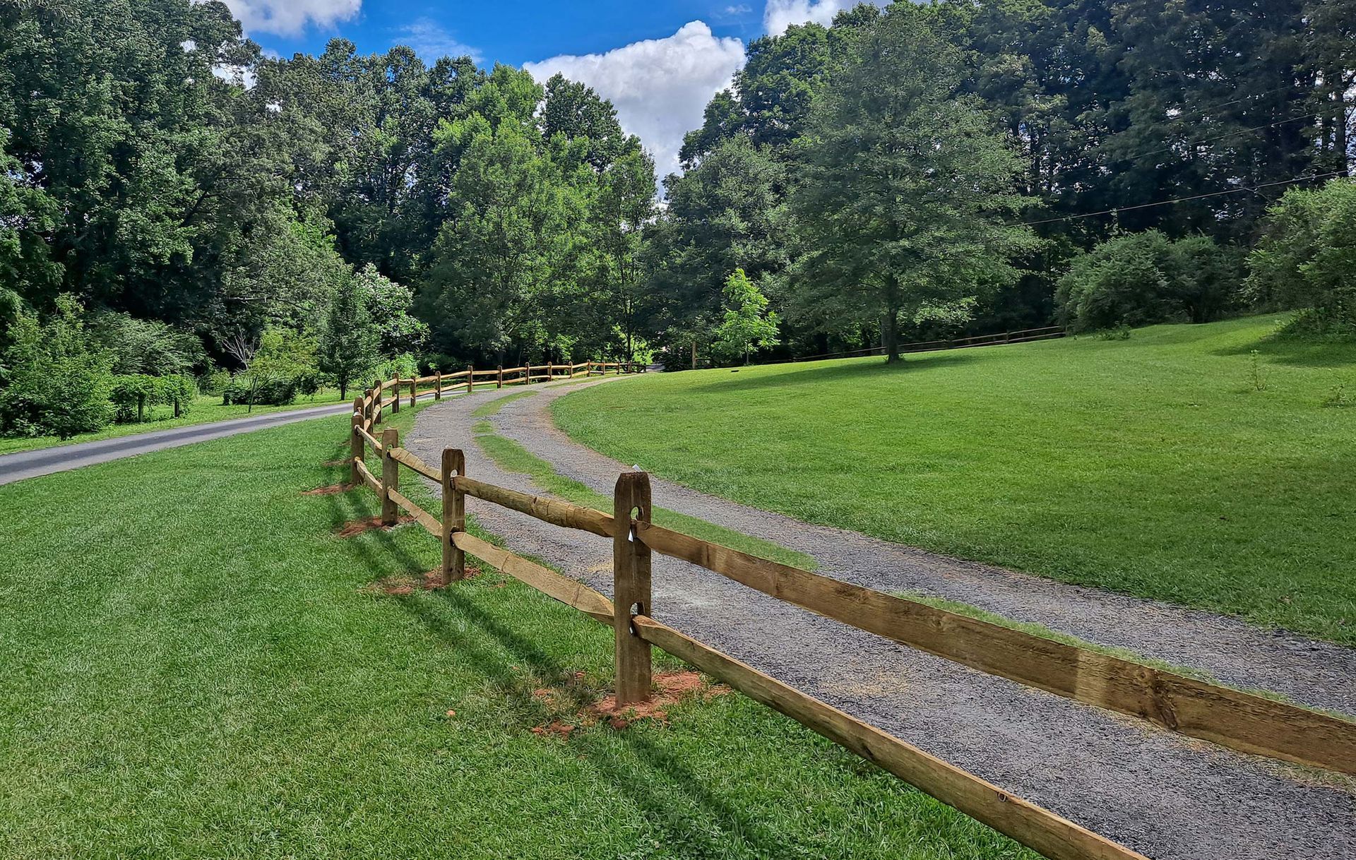 A gravel driveway curves through a grassy field, bordered by a wooden split-rail fence, trees, and blue sky.