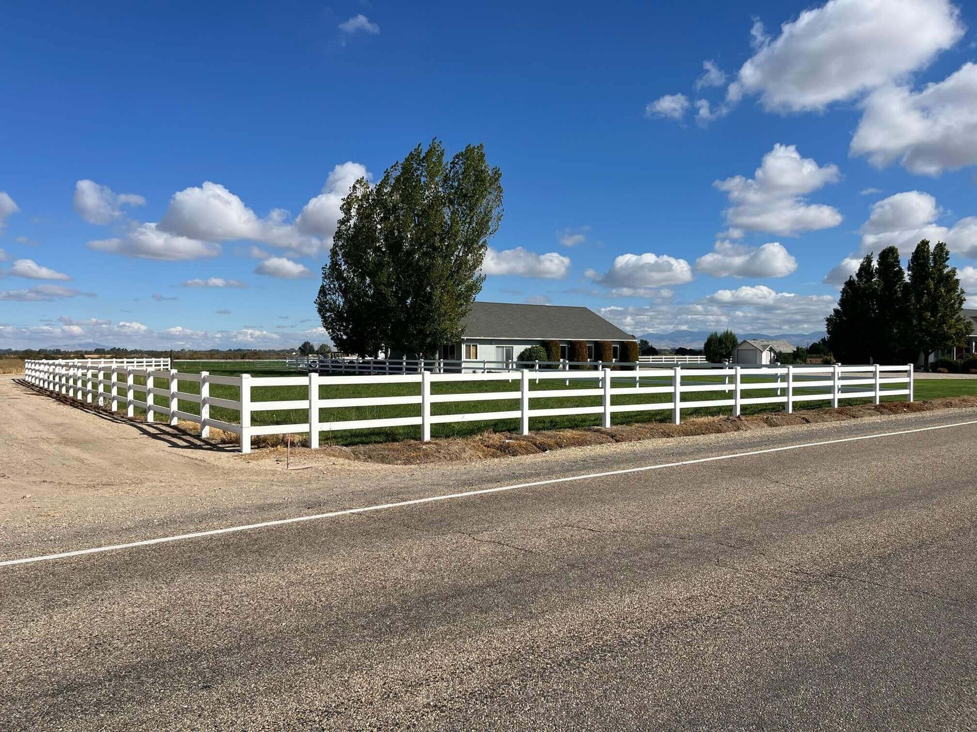 White-fenced house on a gravel road under a blue sky with clouds.