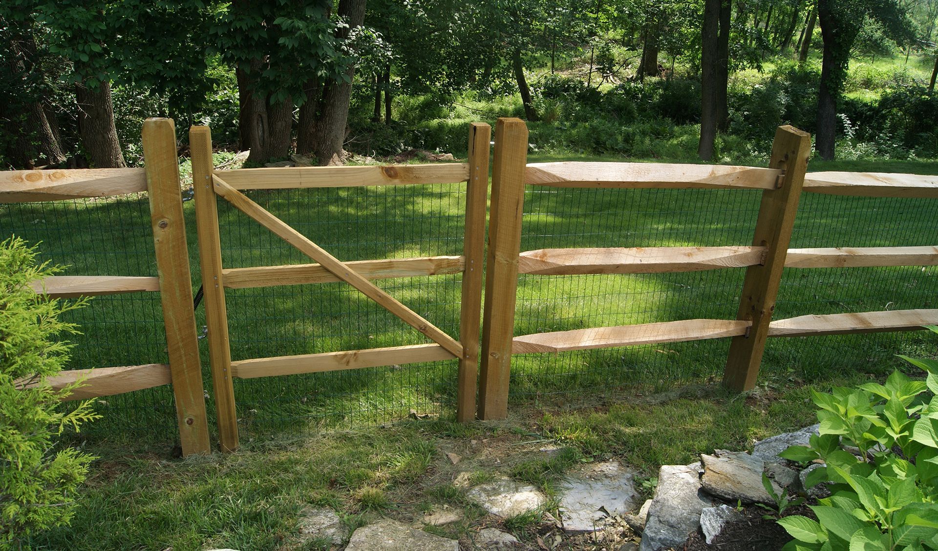 Wooden split-rail fence with gate in a grassy yard, trees in the background, sunny day.