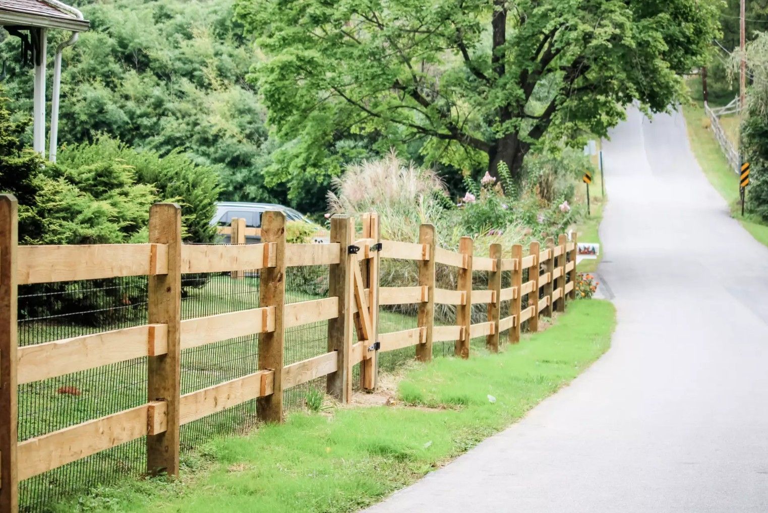 Wooden fence along a road, green grass, trees, and a partly cloudy sky.