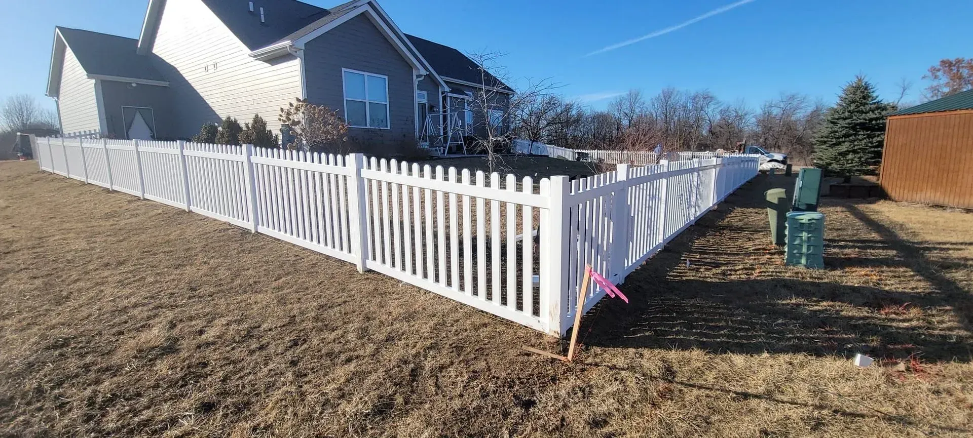 White picket fence around a house on a clear, sunny day. Brown yard and blue sky.