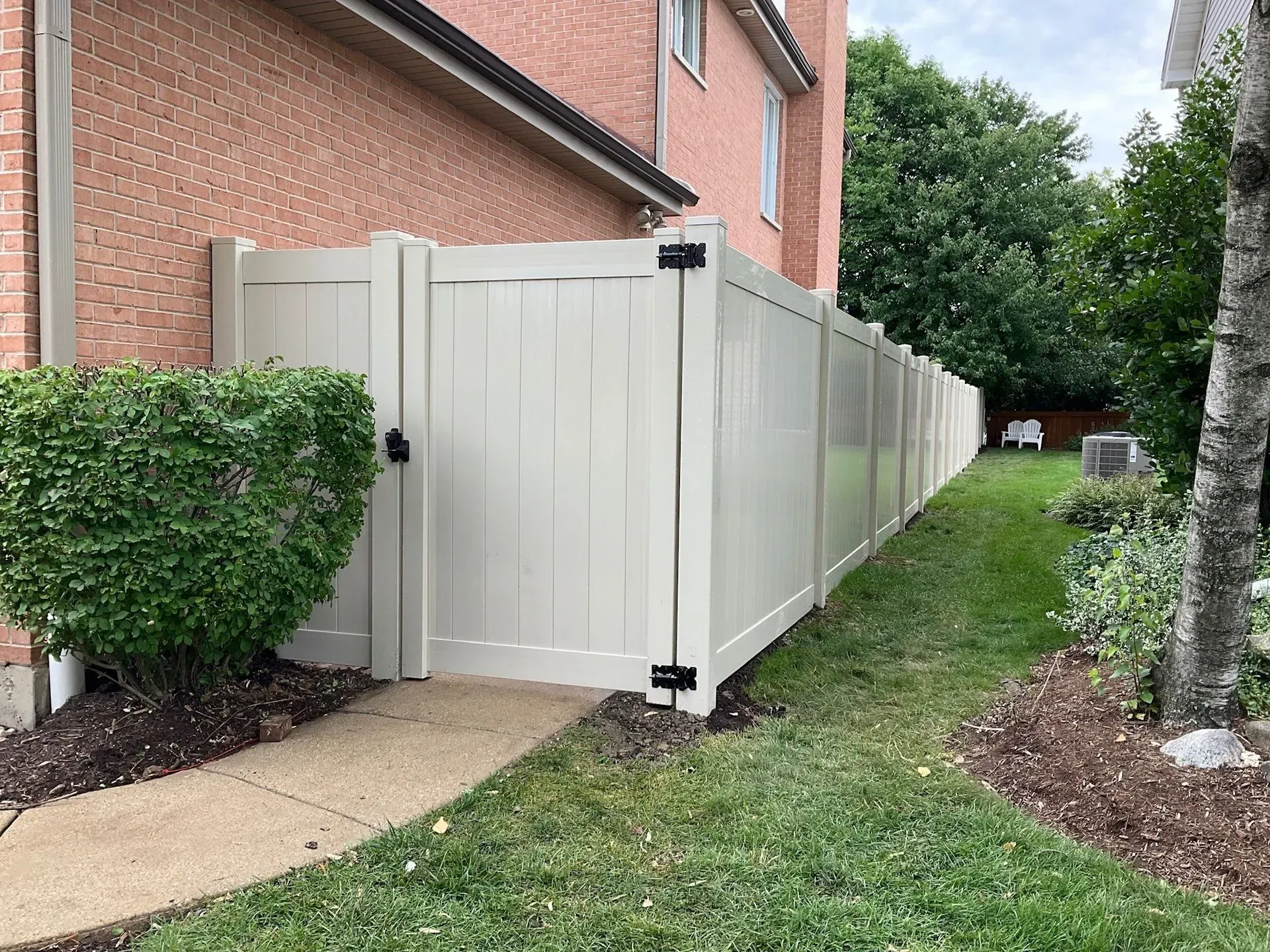 Tan vinyl fence along a brick building, with a gate and landscaping.