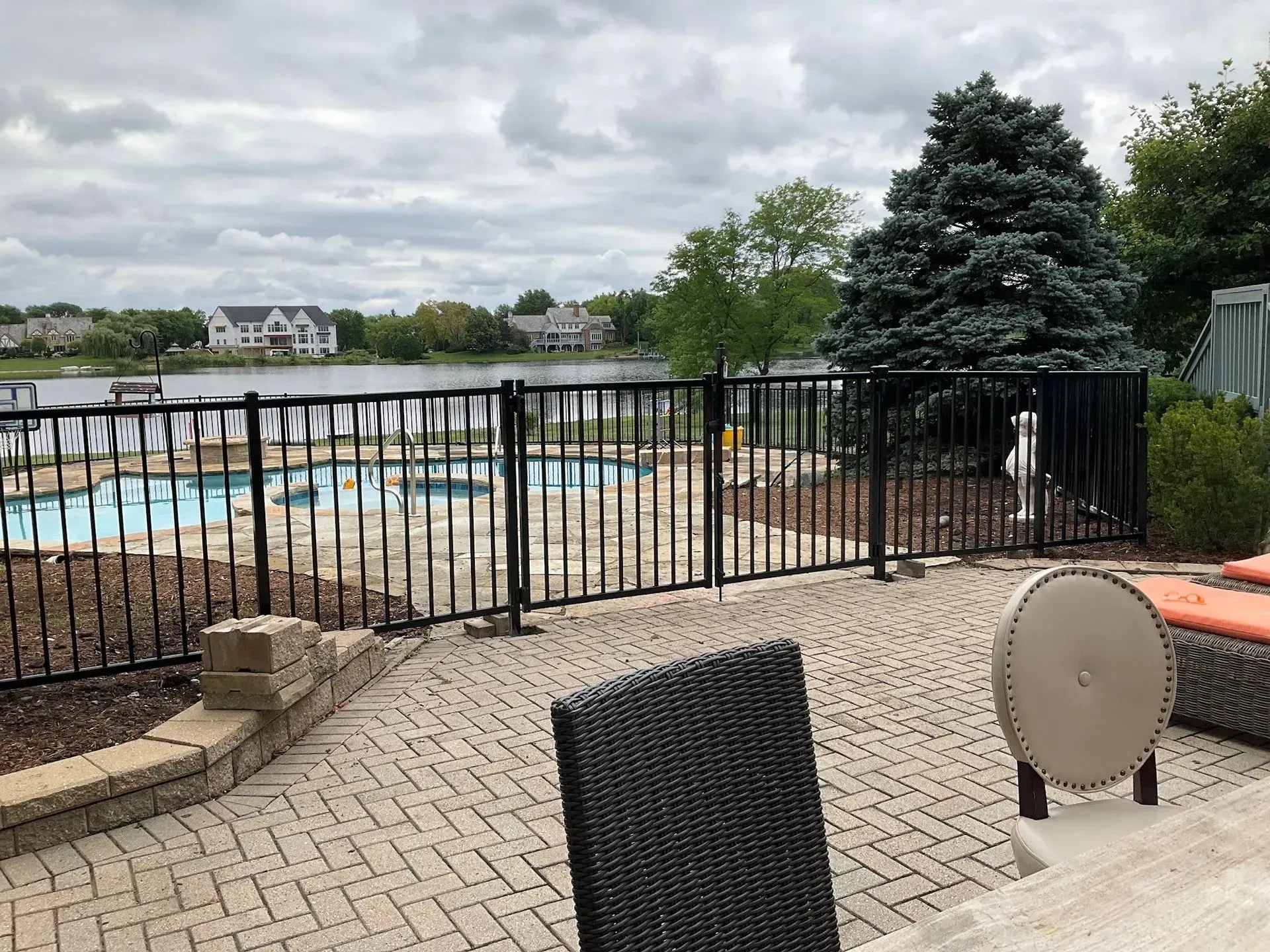 Patio overlooking a pool and lake behind a black metal fence, on a cloudy day.