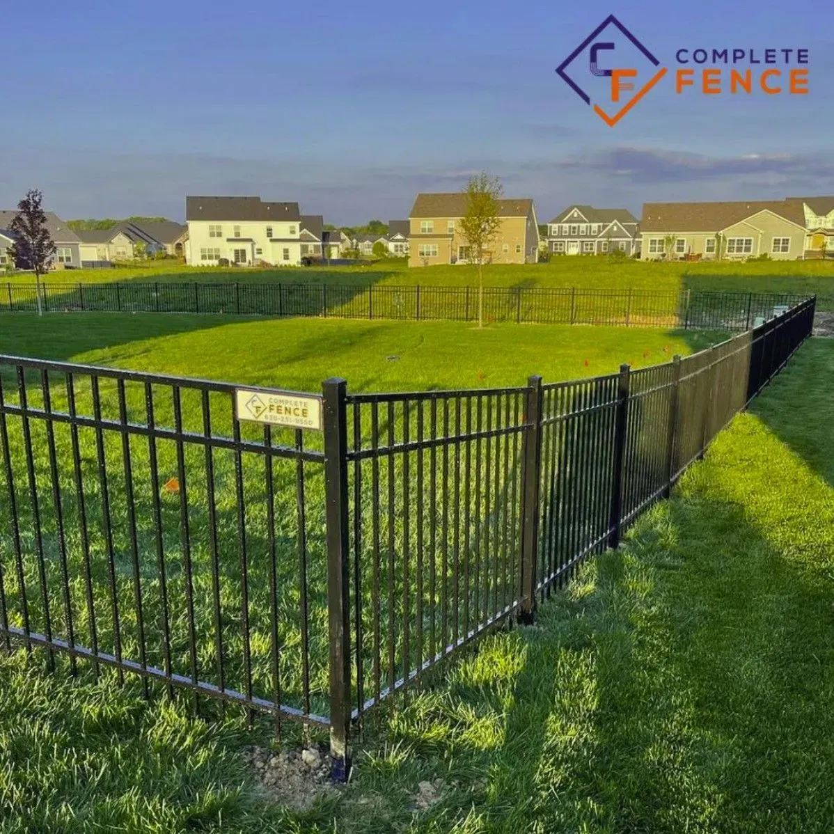Black metal fence surrounding a grassy area with houses in the background under a blue sky.
