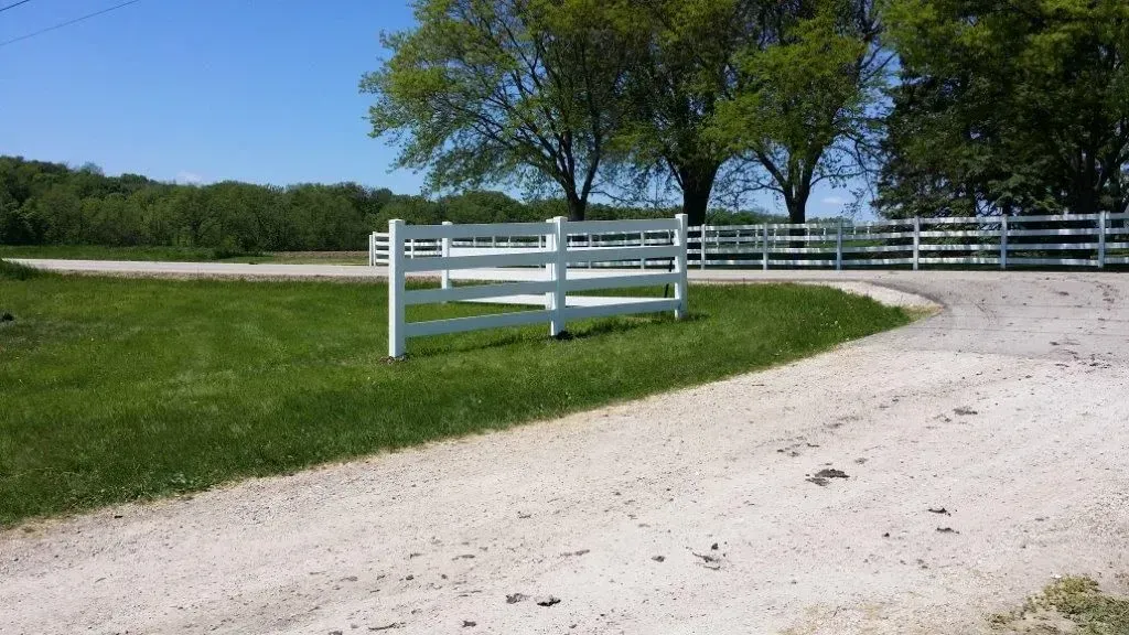 White fence borders a gravel driveway and green lawn, trees in the background under a blue sky.