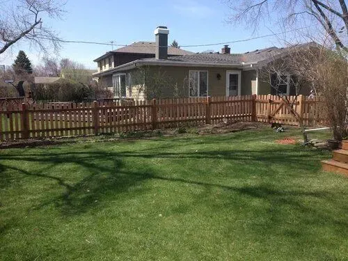 Brown picket fence surrounds a backyard with a house, green grass, and trees.