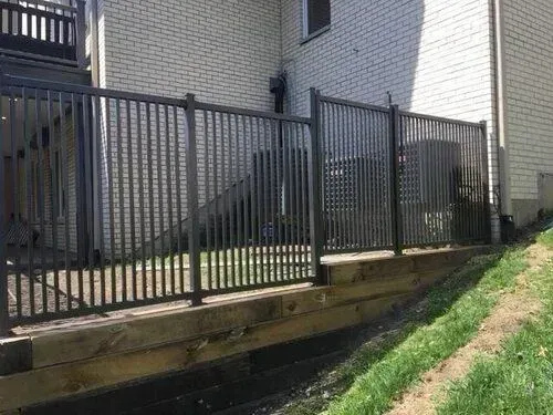 Black metal fence on a wooden retaining wall, with a white brick building in the background and a grassy slope on the right.