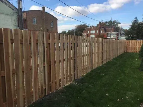 Wooden fence along a grassy yard, with brick buildings in the background under a blue sky.