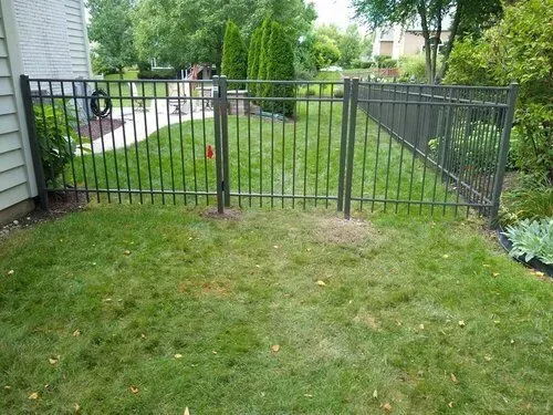 Black metal fence encloses a section of green lawn, with a gate and a house visible.
