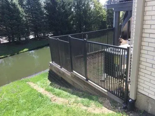 Black fence along water's edge, adjacent to a building with brick and deck, on a sunny day.