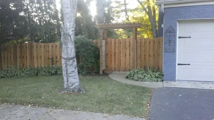 Wooden fence with gate, arbor, and landscaping next to a blue garage.