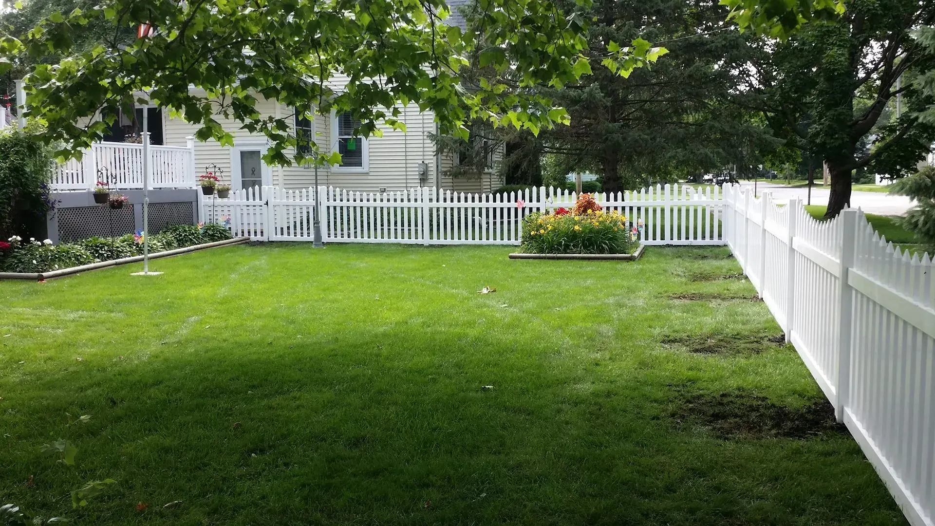 Lawn enclosed by white picket fence, a house, and greenery. Flowers in a raised bed are in the center.