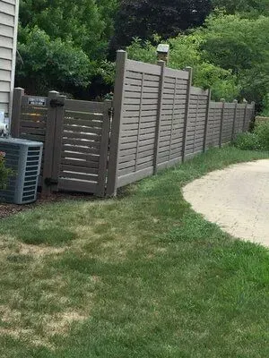 Gray slatted privacy fence with a gate, next to a house and air conditioning unit, on a green lawn.