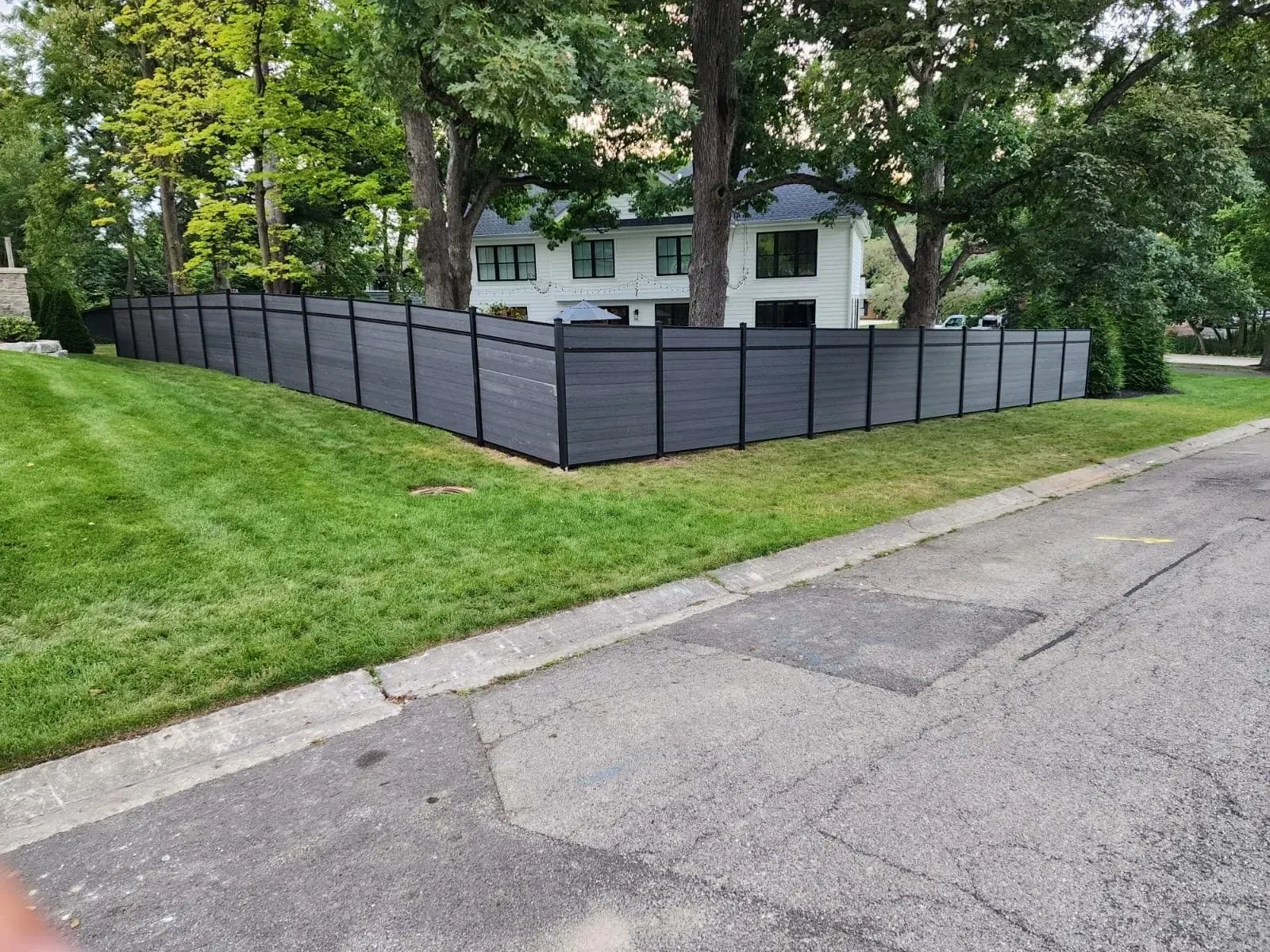 Black fence surrounds a green lawn and white house. Trees flank the property on a street.
