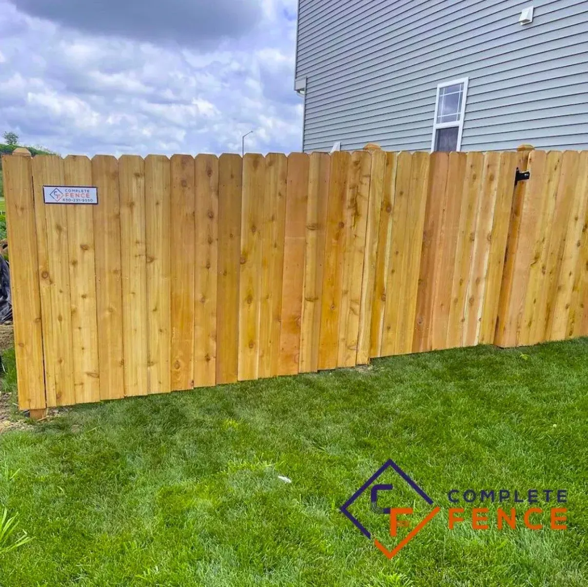 Wooden fence in front of a house, with green grass in the foreground and a cloudy sky.