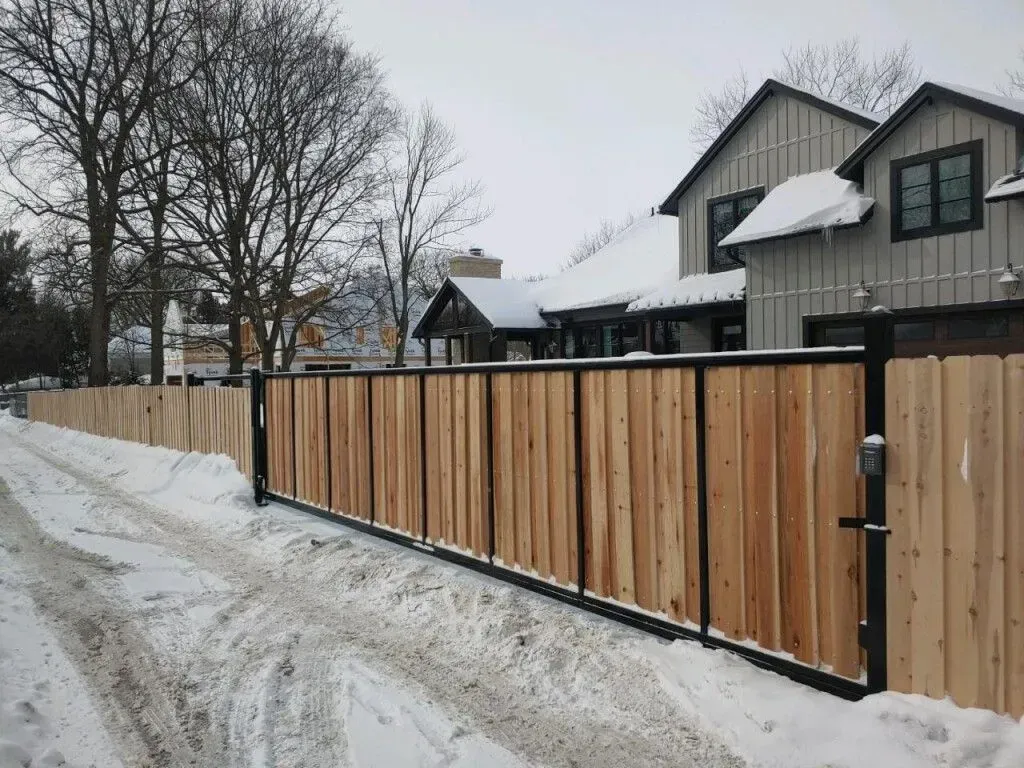Wooden fence with black metal posts in front of a house covered in snow.