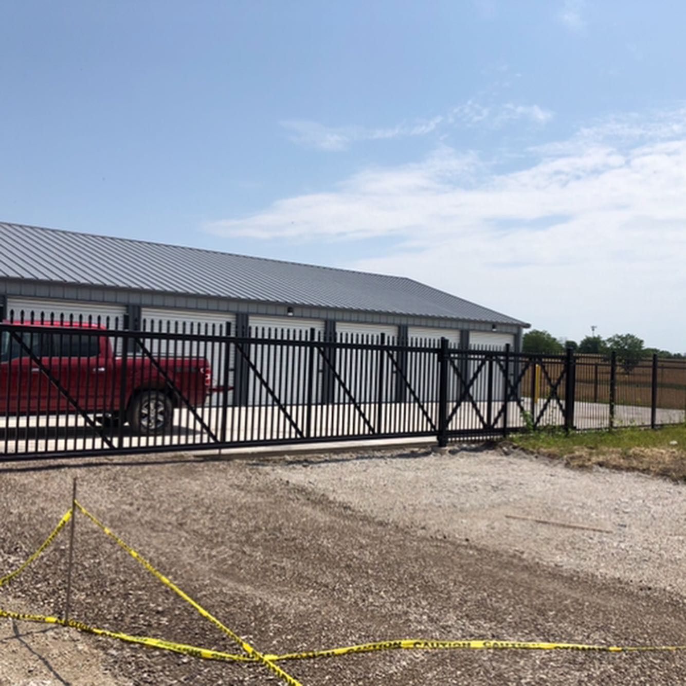 Black gate at storage facility, red truck parked, gravel lot, blue sky.