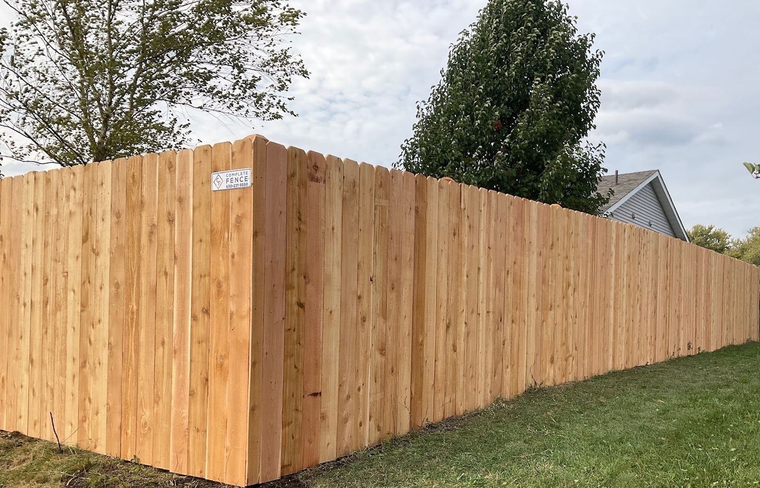 Wooden privacy fence bordering a grassy yard with a house and trees in the background.
