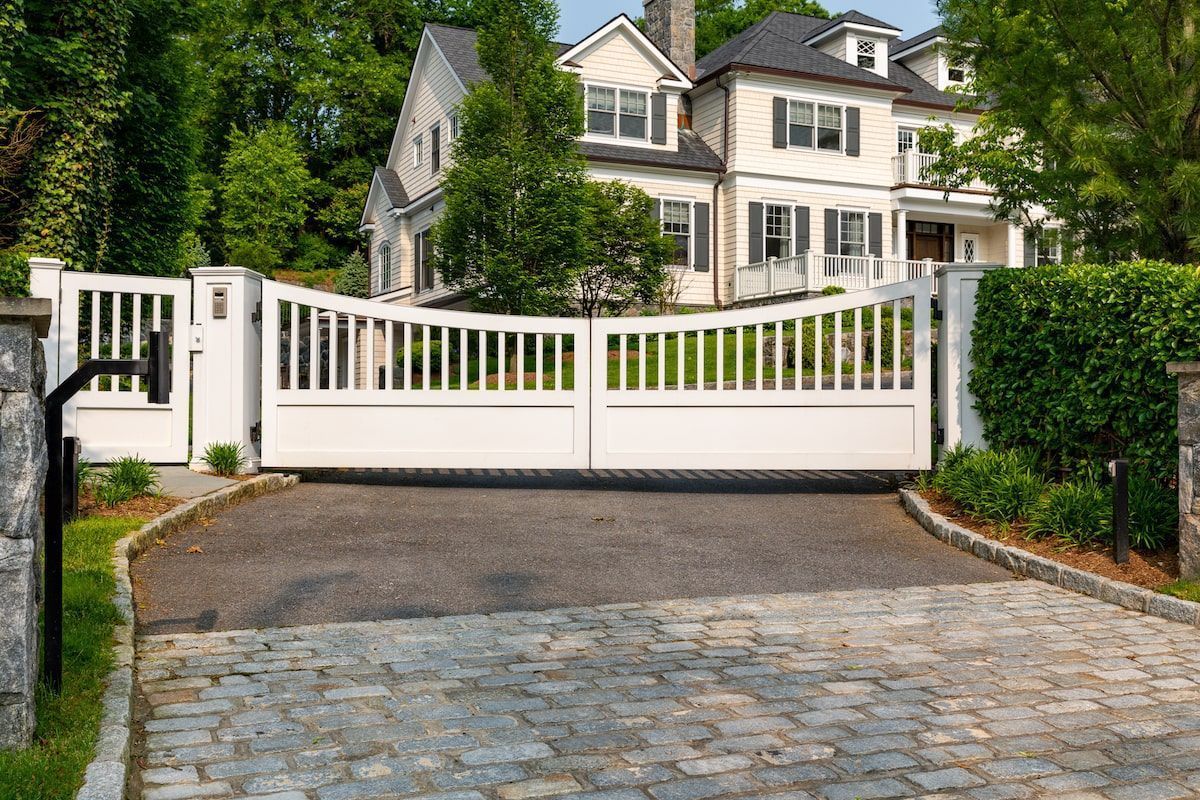 White gated entrance to a large house with a black asphalt driveway and stone accents.