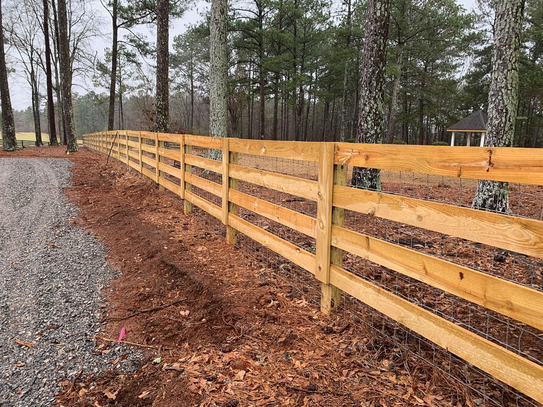 Wooden split-rail fence alongside a gravel driveway and forest, leaves on the ground.