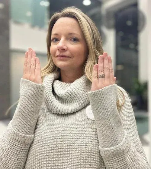 Woman in gray sweater holds hands up, palms out. Light blonde hair, indoor setting.