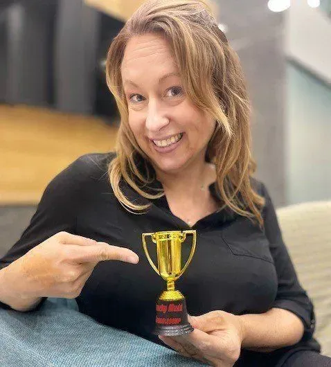 Woman pointing to a small gold trophy; she smiles and sits indoors.