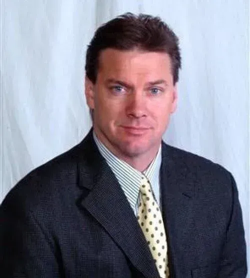 Man in a dark suit with a yellow patterned tie, smiling slightly, against a white backdrop.
