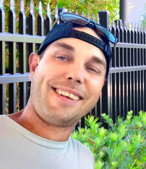 Man wearing a hat and sunglasses smiles near a black fence and green plants.
