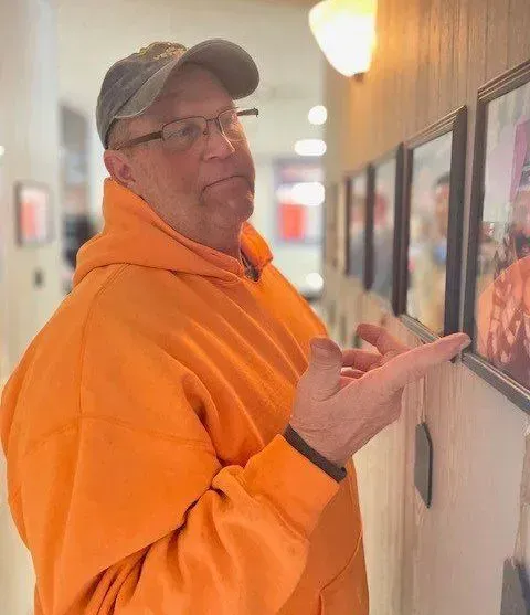 Man in orange hoodie and hat points at framed photographs on a wall.