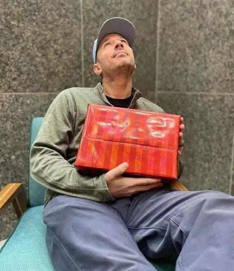 Man in a hat sits, holding a red gift, looking up.