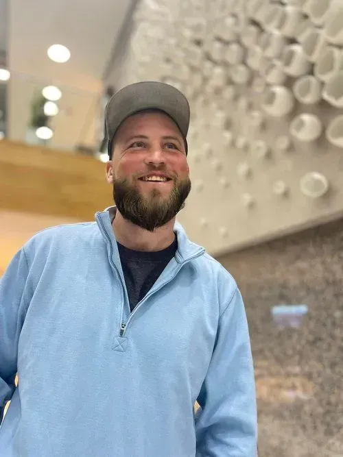 Man with a beard and baseball cap smiles, wearing a light blue pullover in a building with a white art installation.