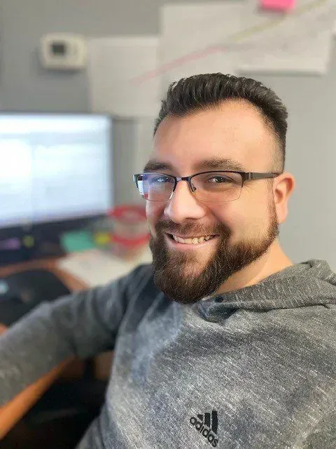 Man with a beard and glasses smiling at the camera, wearing a gray hoodie, in an office setting.