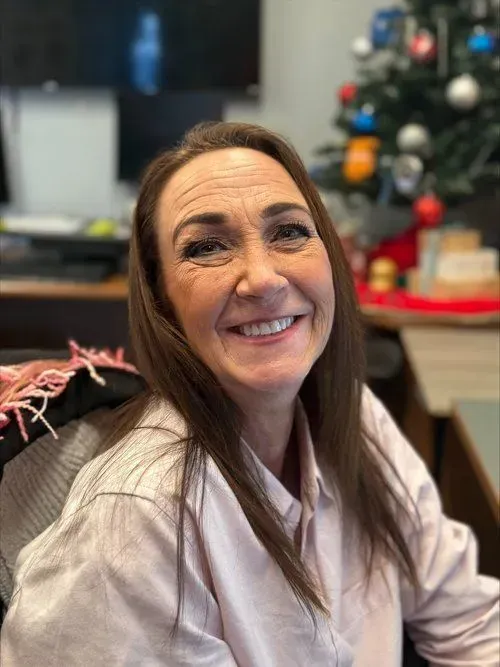 Woman with brown hair smiles, wearing a pink shirt, in an office setting with a Christmas tree.