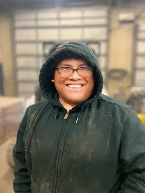 Person in a hooded jacket smiles, wearing glasses.  Indoors, blurred background.