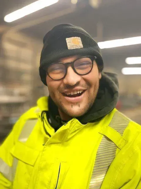 Man in yellow safety vest and black Carhartt beanie smiles, wearing glasses.