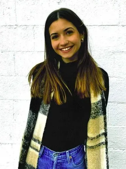 Woman smiling, wearing black turtleneck, blue jeans, and plaid scarf in front of a white brick wall.