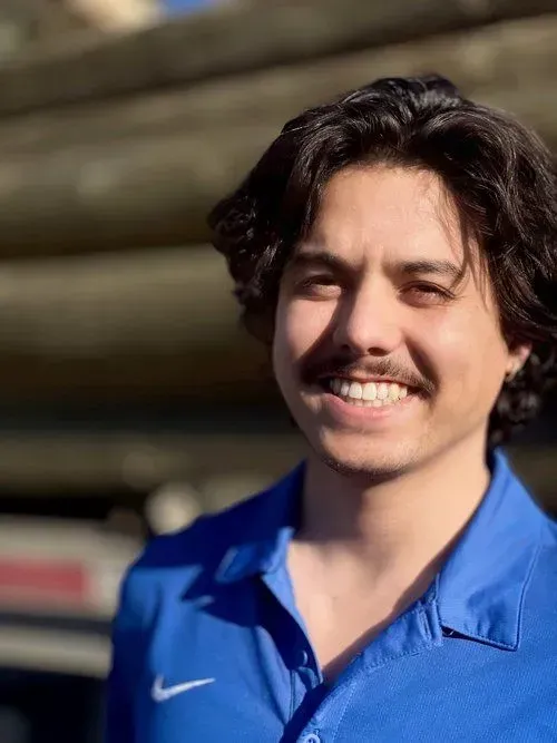 Man with dark hair and mustache smiles, wearing blue shirt in front of wooden background.