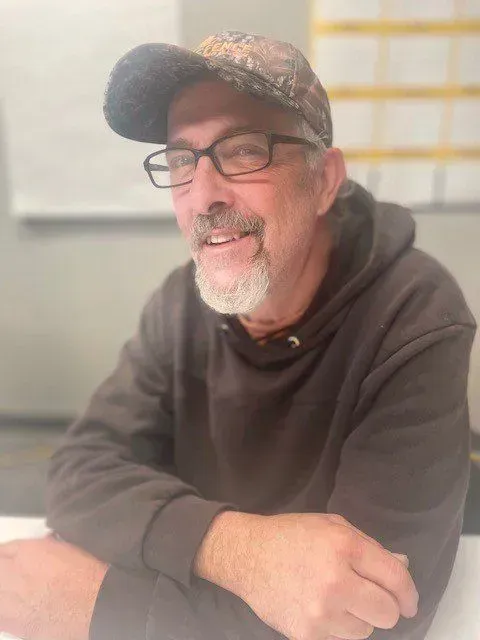 Man wearing glasses and camouflage cap, smiling, sitting at a table, wearing a brown hoodie.