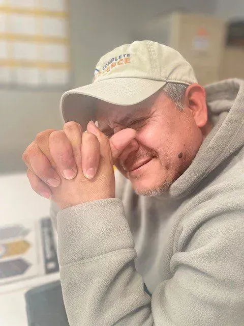 Man in a light-colored hat and hoodie, with hands clasped near face, appears to be praying.