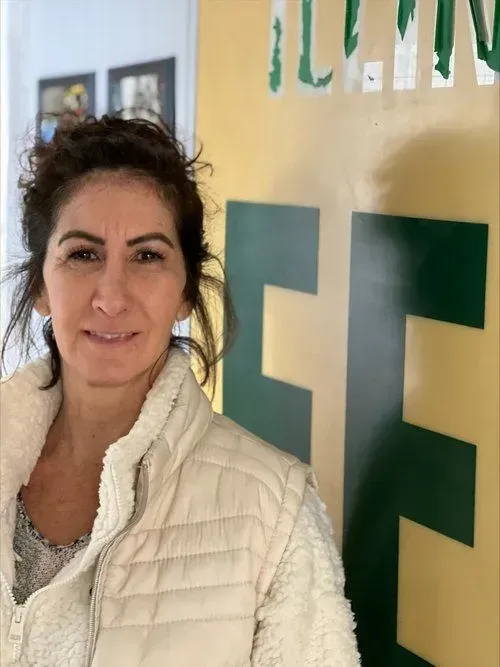 Woman in a cream-colored vest smiles in front of a yellow and green sign with letters.