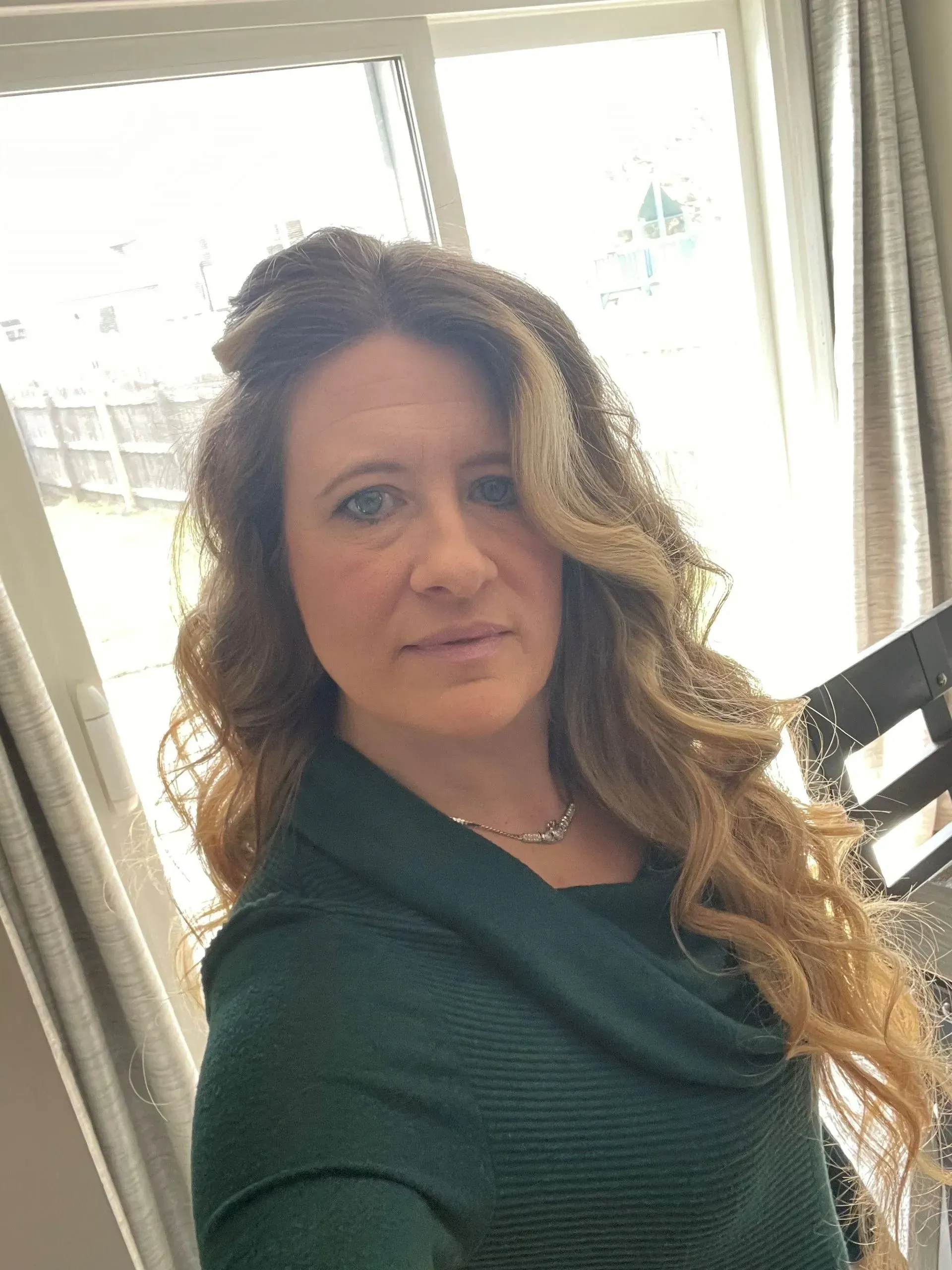 Woman with long wavy hair in a green sweater, looking directly at the camera indoors.