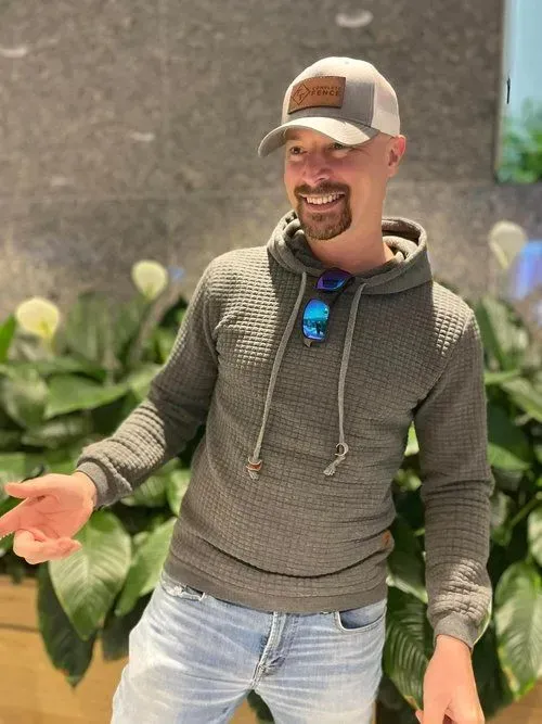 Man smiling, wearing a gray hoodie, baseball cap, and jeans. Standing near plants, gesturing with his hand.