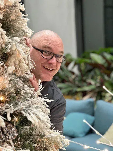 Man peeks around snowy Christmas tree, smiling, wearing glasses.  Blue couch and plants in the background.
