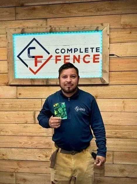 Man holding gift card, standing in front of Complete Fence sign, wooden wall background.