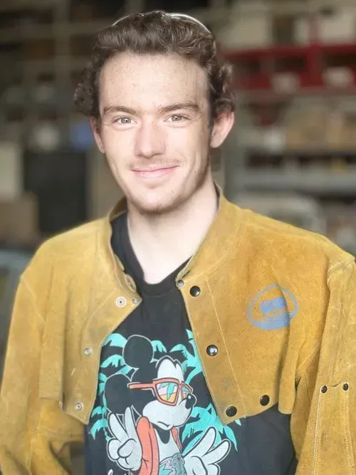 Man smiling, wearing a yellow welding jacket over a black shirt with a Mickey Mouse graphic; indoors.