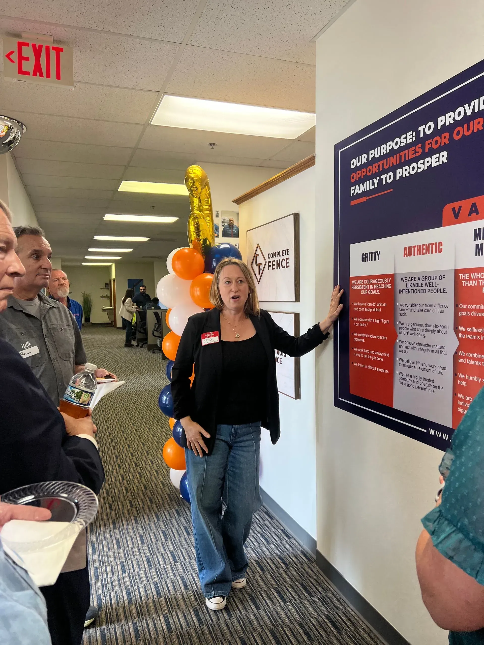 Woman presenting information on a wall poster to a group in an office hallway; balloons and exit sign.